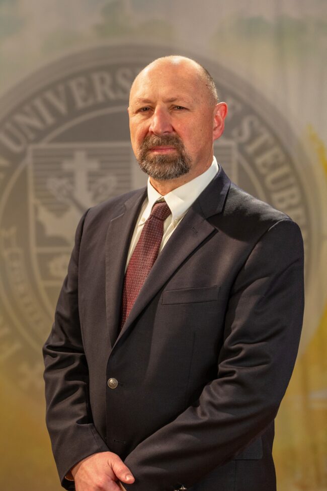 Faculty portrait of Dr. Aaron Urbanczyk standing in front of the Franciscan University of Steubenville seal.