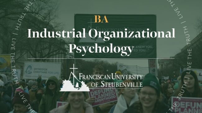 A crowd holding signs at an outdoor gathering with text “BA Industrial Organizational Psychology” and “Franciscan University of Steubenville,” with “Live the Truth” around the border.